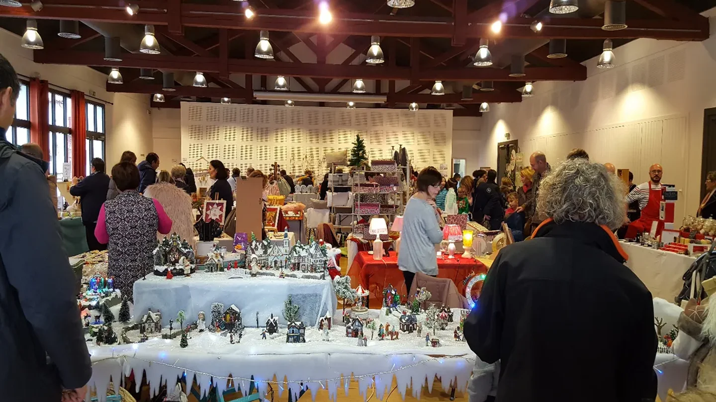 Couple de voyageurs chaudement vêtus tenant une chope de vin chaud fumant devant les façades d’un marché de Noël illuminé